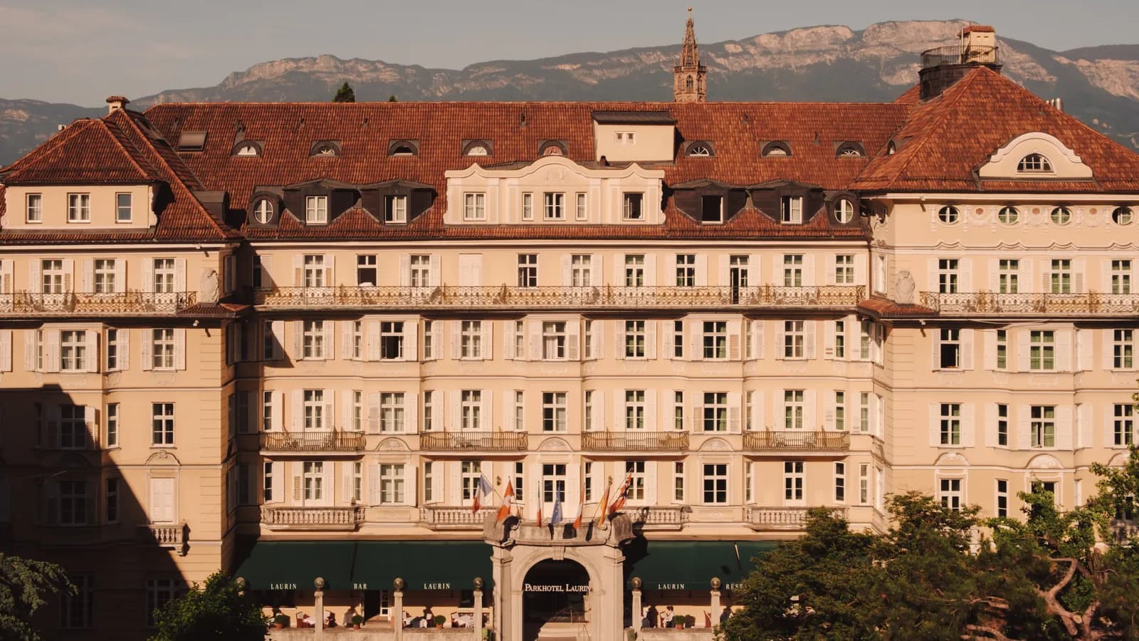 Die Fassade des Parkhotels Laurin in warmem Sonnenlicht, dahinter der Turm des Bozner Doms und die Berge
