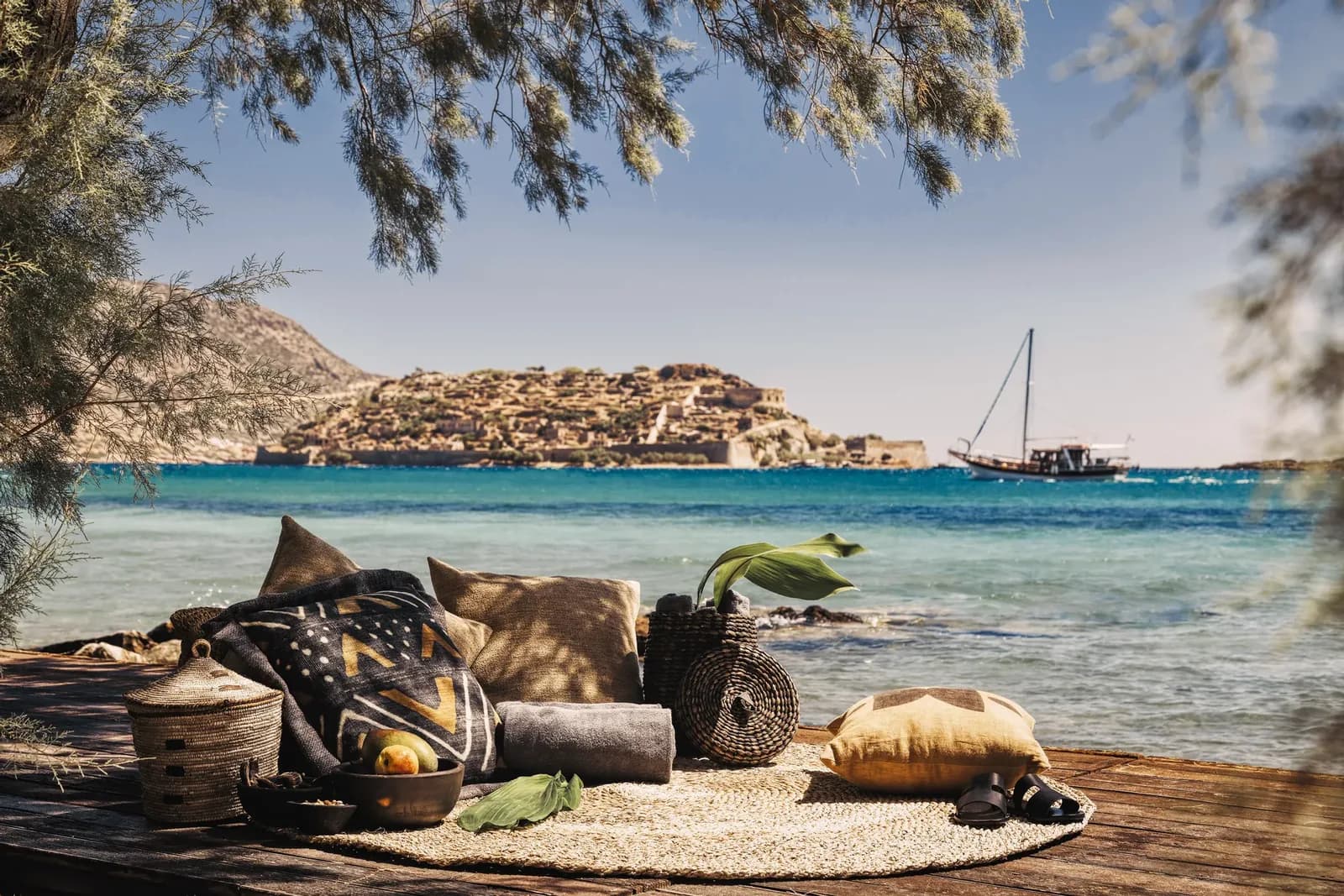 Kissen und Körbe auf einem Holzdeck am Strand von Domes of Elounda, dahinter Spinalonga